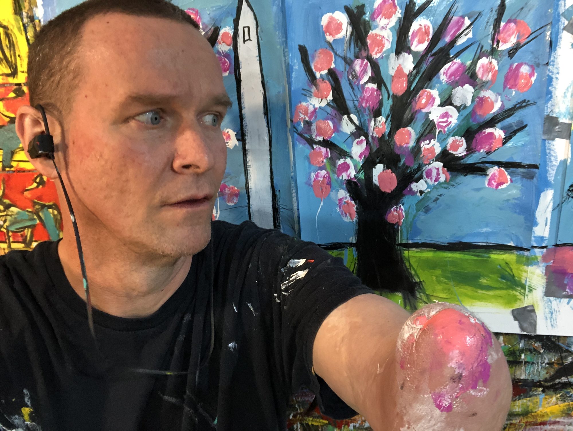 Matt Sesow in his Washington, DC studio — paint-spattered black shirt, earbuds in, his painting arm raised, the tip coated in pink and magenta paint, his bright paintings of the Washington Monument and a blossoming tree behind him